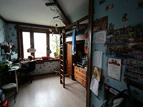 угловой компютерный стол с полками и монитором, лестница на второй этаж с кроватью над столом и шкафом для одежды, многочисленные плакаты на стене детской комнаты простой семейной квартиры