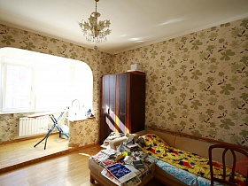 пуфик, заваленный различными вещами у бежевого дивана, шкаф для одежды в спальной комнате, совмещенной с лождией простой большой квартиры в Марьино