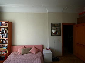 белый торшер, прикроватная тумбочка и книжный шкаф у кровати с розовым вязанным покрывалом в спальной комнате квартиры художника