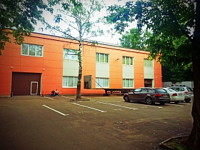 ряд легковых машин во дворе с парковочными местами напротив двухэтажного здания ораньжевого цвета в окружении зеленых деревьев