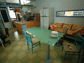 зеленый стеклянный стол и стулья, мягкий диван с подушками в столовой и зона кухни за деревянной стойкой придорожного кафе при гостевом двухэтажном доме