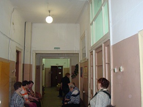 коричневая двустворчатая входная дверь в здание старой действующей больницы из открытых дверных проемом светлого коридора с многочисленными  посетителями на стульях