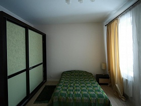 кровать с зеленым покрывалом между шкафом купе и окном в белой спальне