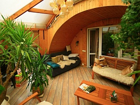 мягкий синий диван с подушками, деревянная мебель и высокие экзотические цветы на просторном застекленном балконе под высоким сводом крыши деревянной семейной  дачи