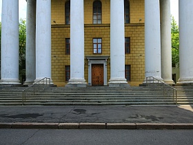 широкое крыльцо усадьбы с металлическими перилами на бетонных ступенях, высокими стройными колонами перед стеной здания, выложенной желтым кирпичом усадьбы советского времени