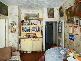 вазы с засушенными цветами на бежевом кухонном шкафу с дверцами , зеркало у входной двери и многочисленные картиры на стене светлой столовой деревянной советской дачи художника с овальной террасой