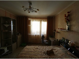 бежевый шкаф под стеклом, открытые боковые полки, комод и столик у окна с коричневыми шторами в спальне двухкомнатной квартиры новостроя