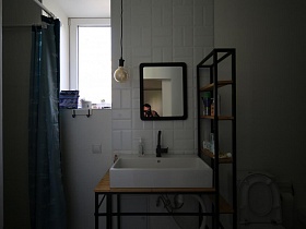 открытый стеллаж с туалетными принадлежностями на деревянных полках между санузлом и белой раковиной на деревянном столике, зеркало в темной рамке над белой раковиной с черным краном, голубая штора, отделяющая душ в ванной комнате современной скандинавско