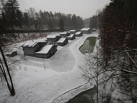 ряд двухэтажных элитных домиков в окружении снега на берегу реки в лесополосе