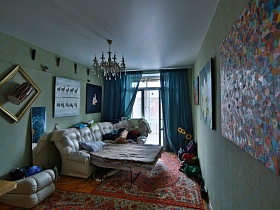 люстра, стилизованная под свечи на белом потолке спальной комнаты с большим угловым разложенным диваном, мозаичной картиной и другими художественными произведениями на салатовых стенах современной квартиры художника
