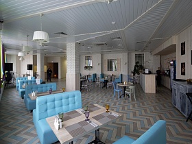 голубые мягкие диваны вокруг прямоугольных столиков в индивидуальной зоне, стулья со спинкой вокруг круглых столиков по центру светлого зала Бар Лофта в спальном районе