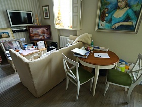 белые деревянные стулья с зелеными седушками у круглого деревянного стола на фигурных ножках, белый мягкий диван с подушками в гостинной с многочисленными картинами в рамках девчачьей дизайнерской квартиры
