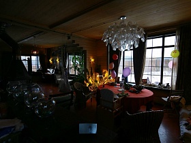 большая белая люстра на деревянном потолке над обеденным круглым столом с красной скатертью гостиной с коричневыми шторами на больших окнах уютной избы