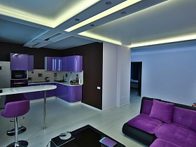белый барный столик на металлической ножке на границе кухни с фиолетовой мебельной стенкой у коричневой стены и гостиной с яркими фиолетовыми подушками на угловом мягком диване