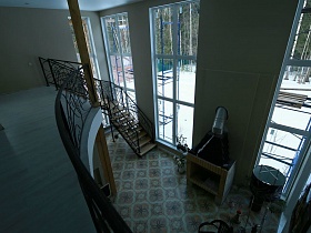 высокие окна в два этажа просторной светлой гостиной с кирпичным камином у стены между окнами и деревянной лестницей с кованными фигурными перилами недостроенного элитного дома