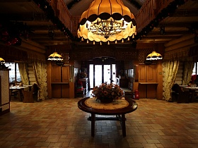 большая люстра, стилизованная под свечи под желтым абажуром над круглым деревянным столом с цветами в центре просторного холла напротив входных дверей купеческого ресторана