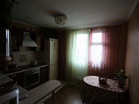 барная стойка у коричневой мебельной стенки, белый двухкамерный холодильник и обеденный стол с цветной скатертью на кухне у окна с балконной дверью