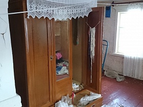 открытый трехдверный деревянный шкаф с зеркалом посередине с вещами в захламленной комнате дома заброшенной деревни