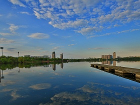 выступающий длинный деревянный пирс на бетонных опорах чистого озера с зеркальным отражением белых облаков и синего неба