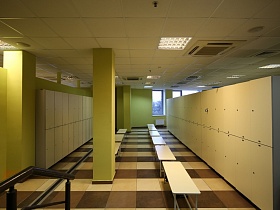 просторный зал раздевалки с белыми шкафами у салатовых стен с колоннами, бежево коричневой плиткой на полу , освещением и кондиционерами в потолке провинциального фитнеса