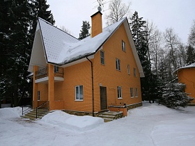 общий вид кирпичного двухэтажного дома с балконом,лестницей с перилами между двух колонн у крыльца на зимнем участке