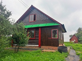 коричневый деревянный домик с треугольной крышей  на участке с зеленой травой, деревьями большой круглой бочкой у тропинки
