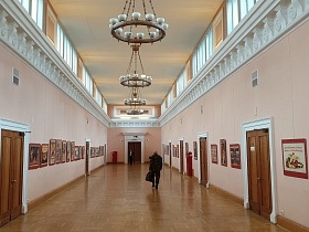 просторный высокий зал галереи с большими окнами под потолком, круглыми подвесными круглыми люстрами на цепях с белыми плафонами и многочисленными историческими плакатами в рамках на светлых стенах эпохи СССР