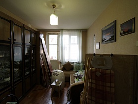 картины на стене над бежевым угловым мягким диваном с подушками у окна с балконной дверью, многочисленные доски, гладильная доска, табурет, мобильный кондиционер у мебельной стенки с посудой в гостиной простой квартиры