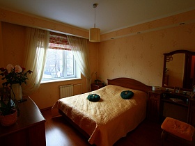 круглые зеленые декоративные подушки на деревянной кровати с бежевым стеганным покрывалом, зеркало над коричневым гримерным столиком в спальной комнате квартиры государственного служащего