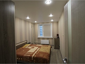 большая кровать с коричневым постельным в светло серой спальне двухкомнатной квартиры высотного здания в новострое