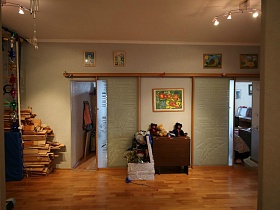 доски в углу за спортвным уголком,складной стол с игрушками у стены, между двумя смежными комнатами,детские картины на светлой стене просторного холла современной трехкомнатной квартиры с ораньжевой кухней
