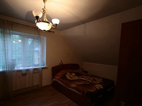 коричневая кровать с цветным покрывалом у окна с прозрачной гардиной, белая люстра на потолке спальни на мансарде семейного двухэтажного дома в густом лесу