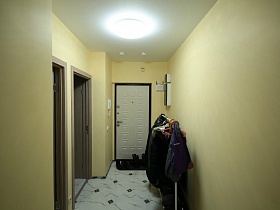 напольная вешалка с одеждой, коврик с обувью у белой входной двери в светлой прихожей молодежной евро квартиры в Новострое