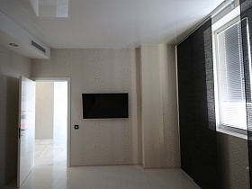 плоский телевизор, черный выключатель на молочного цвета стене спальной комнаты с белым потолком и черными вертикальными жалюзи на большом окне