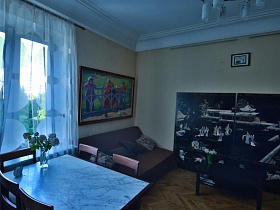 стулья со спинками вокруг прямоуголного светлого мраморного стола у окна с белой гарлиной, коричневый мягкий диван с подушками, картины на столе и на полу в гостиной стильной квартиры художника