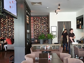 серые деревянные стеллажи с бутылками на полках у стены зонированного зала ресторана с уютными сервированными столиками
