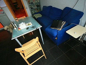 металлический термос, салфетки, кружка на прямоуголном столе, небольшой белый складной столик,ноутбук на подушке синего мягкого дивана на полу с темно серой плиткой кухни семейной трехкомнатной квартиры