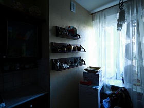 декоративная тарелка, посуда, и статуэтки на открытых деревянных полках на стене у окна с белой гардиной  в зоне кухни квартиры художника