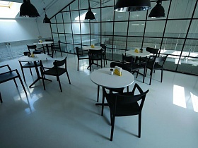 черные пластиковые кресла вокруг белых круглых столиков на черных ножках на белом полу уютного кафе лофт с прозрачной стеклянной стеной