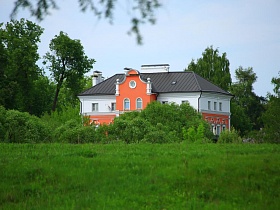 необычное двухцветное двухэтажное здание с ораньжевым фасадом и серой крышей среди густой зелени деревьев и густой травы в живописном месте Подмосковья