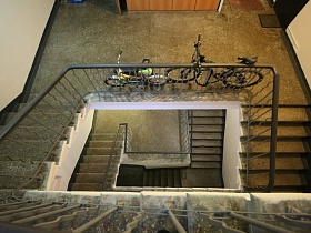 два велосипеда у металлических перил на площадке этажа 9 подъезда с лестничными бетонными маршами
