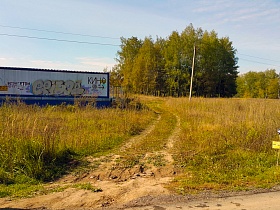 высокая трава прикрывает огромный рекламный щит на металлическом сооружении у развилки дорог в поле
