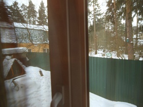 вид из окна сквозь белую гардину на заснеженный участок съемного дома за зеленым забором в сосновом лесу