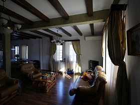общий вид гостиной с мягкой мебелью, телевизором, подвесными люстрами на белом потолке с деревянными балками для съемок кино в зонированной комнате