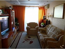 светлый ковер с цветком между рыжей стенкой и белым мягким диваном с подушками в гостиной с рыжими шторами на окне  трехкомнатной квартиры панельного дома
