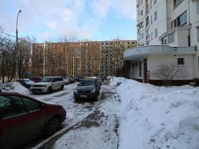 расчищенный от снега пешеходный тротуар и дорога с припаркованными машинами напротив подъезда жилого дома с полукруглым навесом