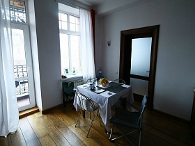 металлические стулья вокруг квадратного обеденного стола с серыми индивидуальными салфетками на белой скатерти в кухне с белой гардиной на окне и балконной двери