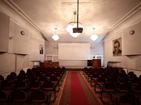 большие подвесные люстры на цепях на белом потолке просторного актового зала с красной дорожкой между красными секционными креслами на паркетном полу и портретами известных людей на стенах ДК эпохи СССР