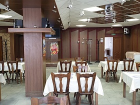 коричневые деревянные стулья со спинкой вокруг столов с белой скатертью по периметру просторного зала столовой с панельными стенами,квадратными под дерево колонами и зеркальным потолком