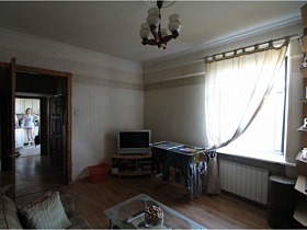 телевизор на тумбе в углу гостиной, сушилка с бельем у окна с белой гардиной, стеклянный столик у дивана, подвесная люстра с круглыми плафонами на белом потолке простой сталинской квартиры 90-ых для съемок кино